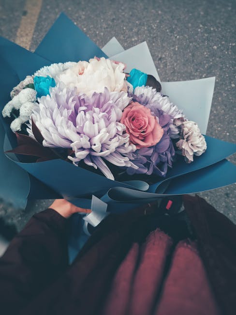 A hand holding a vibrant floral bouquet arranged with white chrysanthemums, light purple chrysanthemums, a pink rose, and small blue accents, wrapped in dark blue paper with light blue and white layer