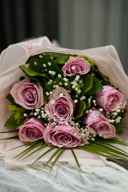 A beautifully arranged bouquet of fresh pink roses with full, velvety petals and lush green leaves, complemented by small white baby's breath flowers. The roses are wrapped in elegant white tissue pap
