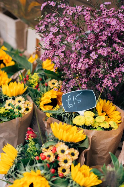 A vibrant display of fresh floral bouquets arranged at a flower market stall, featuring bright yellow sunflowers, small white daisies with yellow centers, and red and pink flowers in brown paper wrapp