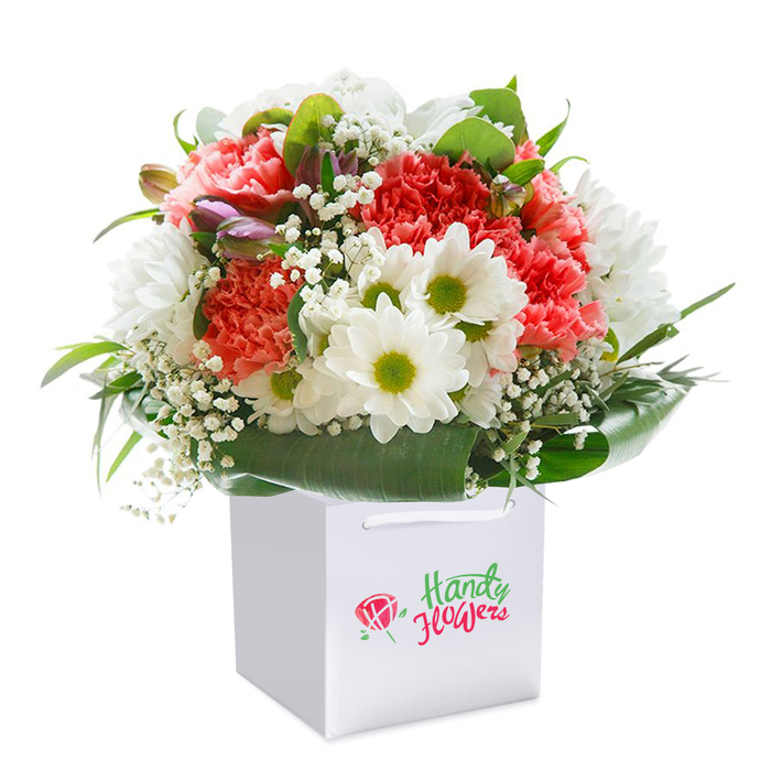 Poised in a sleek, square white bag, this vibrant floral bouquet is a testament to the detail-oriented floristry found in Forest Gate. The arrangement harmoniously blends the lush, ruffled petals of coral, peach, and ruby-red carnations-each one bursting with color and gentle fragrance-making for an inviting focal point. Dotted throughout are crisp white daisies, their bold yellow-green centers radiating optimism, much like the spirit of bustling Woodgrange Road. Wisps of dainty baby's breath add lightness and texture, their tiny blossoms evoking the soft mist of a cool morning in Plashet Park. Around the base, broad, dark emerald leaves splay outward in graceful arcs, forming a natural collar that both protects and elevates the intricate blooms above. Subtle hints of closed, purple-tinged buds peek from the side, lending mystery and serene contrast within the arrangement. The bouquet's full, rounded silhouette and careful layering of color and texture are highlighted by the bag's sturdy ribbon handles, designed for easy carrying through Forest Gate's leafy avenues. Set against a flawless white background, the verdant greenery and radiant flowers are given space to shine, each detail thoughtfully curated for elegance and delight. Perfect as a gesture for life's joyful milestones or to send a heartfelt message, this piece reflects the freshness and artistry of Garden City's finest florists and promises swift, reliable delivery throughout the local area.