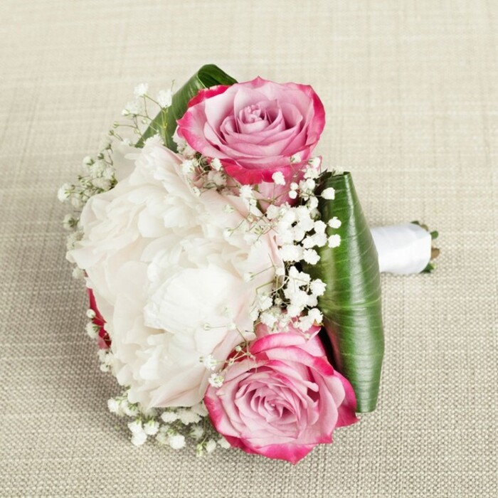 Expertly crafted by the florists at Flower Delivery Forest Gate, this lavish bridesmaid's bouquet rests upon a lightly textured, neutral fabric, evoking the understated charm of a Forest Gate afternoon gathering. Dominating the arrangement, a sumptuous white peony blooms in the centre, its plentifully layered, ruffled petals forming a creamy, cushion-like focal point that invites the eye. On either side, two pink roses elegantly anchor the design - their outer petals tinged with vivid fuchsia before fading into soft blush and faint lavender at the inner spiral, symbolizing heartfelt romance and graceful transformation. Throughout the bouquet, finely interwoven sprigs of baby's breath (Gypsophila) add a whimsical, snowflake-like delicacy, lending the piece a lightness as enchanting as the first days of spring in West Ham Park. The bouquet's base features luxuriously folded emerald-green leaves, offering a vibrant, tailored structure and subtle shine against the flowers' gentle hues. The stems are unified by a chic white satin ribbon handle, finishing the composition with poise and clarity - easy to grip for processions through Forest Gate's historic avenues or festive gatherings at local venues. The lighting brings out each intricate petal and the crispness of the leaves, highlighting the attention to detail characteristic of local floral designers. This floral creation is perfect for weddings, milestone celebrations, or as a classic gift, exuding elegance, freshness, and the welcoming spirit of Forest Gate.