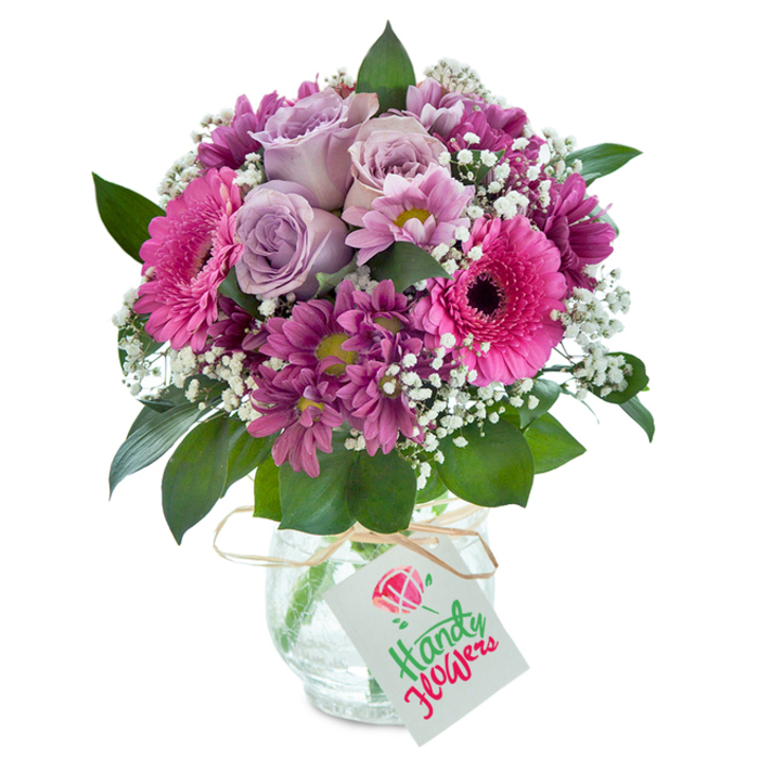 Taking centre stage against a luminous white background, this vibrant mixed bouquet radiates colour and expert craftsmanship-reflecting the lively spirit of Forest Gate's neighbourhood florists. Soft lavender roses, with petals unfolding gracefully, form the inviting heart of the arrangement, bringing both visual calm and the suggestion of sweet, gentle fragrance. Encircling them are bold magenta gerbera daisies, their wide petals and dark eyes brightening the bouquet with confident cheer, reminiscent of the vivid floral displays seen near Romford Road's bustling cafés. Contributing to the arrangement's rich complexity, clusters of deep plum chrysanthemums add texture and a sense of depth, with subtle yellow-green centers that play with the natural light, reminiscent of the overarching shade in Forest Lane Park. Delicate sprigs of baby's breath create a gentle, cloud-like softness, while abundant dark green leaves peek from beneath, providing a lush, natural foundation that harks back to Forest Gate's green spaces. The bouquet is showcased in a clear, crackle-glass vase that catches the light with understated elegance, tied at the neck with rustic raffia twine for a homespun touch. From the twine, a rectangular white gift tag dangles, featuring a joyful birthday message in red and green lettering, just above a miniature flower illustration. This thoughtfully curated bouquet makes an ideal gift for birthdays, anniversaries, or moments of gratitude, especially for residents of Forest Gate and the wider E7 area, embodying the artistry, vibrance and personal attention local florists bring to every heartfelt delivery.