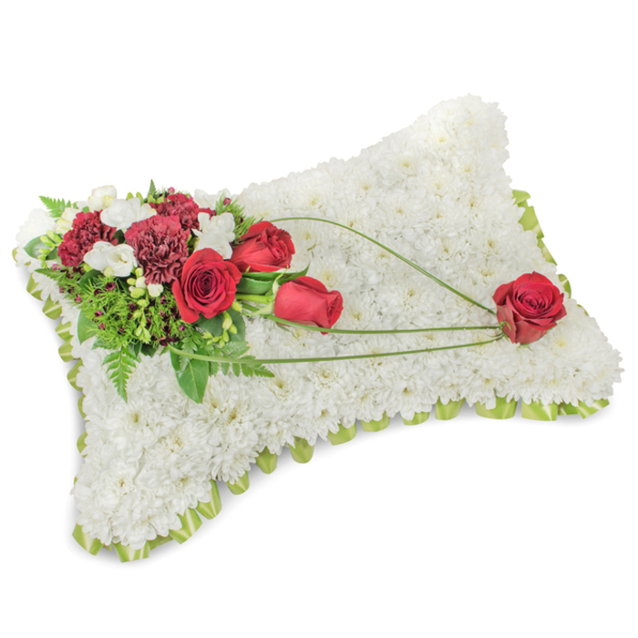 A meticulously assembled memorial pillow, rectangular and gently domed, whose entire visible surface is composed of tightly packed white chrysanthemum heads forming a soft, velour-like texture that suggests peace and repose. The chrysanthemum blanket sits within a subtle outline of light green ribbon or foliage, which peeks out around the edges like a delicate frame. Anchoring the design in the upper left is an abundant spray of deep red flowers-several richly hued roses at different stages of opening paired with deep burgundy carnations-whose saturated tones create a dignified contrast against the snow-white base. Fine clusters of small white blooms, possibly freesias, ripple through the red cluster and temper the palette with gentle highlights, while layers of greenery, including slender fern fronds and rounded leaves, build luminous shadows and depth. From this primary bouquet a trio of slender, glossy green blades or bear-grass arcs across the surface in a graceful diagonal, the sweep culminating in a single, perfectly shaped red rose placed on the lower right quadrant as if gently laid down. The flowing lines of foliage give the piece movement and narrative, leading the eye from cluster to solitary bloom and reinforcing the theme of remembrance. Under soft studio or morning light the arrangement reads as serene and heartfelt-ideal for formal funeral displays, cemetery tributes, or intimate chapel services in Forest Gate and nearby community spaces-evoking comfort, respect, and enduring love without excess.