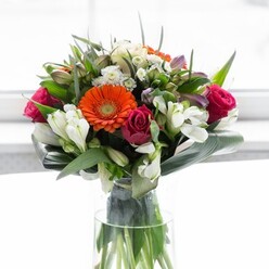 This sophisticated bouquet is a feast for the senses, artfully combining bold colours and delicate textures in a way that captures the unique charm of Forest Gate floristry. The arrangement spotlights plush gerbera daisies-two vivid orange, two peach with creamy undertones-whose soft petals radiate outwards, inviting touch and admiration. Nestled amongst these are rich fuchsia roses, their tightly furled blossoms adding depth and a classic touch of romance. A scattering of white alstroemeria and spray roses punctuates the bouquet with light, airy notes, while softer pinks and tiny white blooms provide subtle highlights that shimmer beneath the overhead light, akin to daylight filtering through the leafy streets near Wanstead Park. The foundational layer of large, curved aspidistra leaves creates a lush emerald collar, grounding the exuberant colour palette and expertly containing the display's volume. Smaller, pointed greenery adds further structure, filling in any gaps and giving the arrangement an abundant, just-picked feel. All these elements are showcased in a slender clear glass vase through which vibrant stems are neatly aligned, signalling freshness and professional care. The soft, diffused background lets the flower colours sing while the composition's warmth and optimism make it perfect for gatherings, celebrations, or as a heartfelt gift for anyone in the vibrant Forest Gate community.